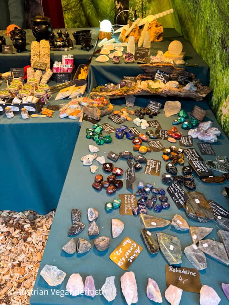 A festive market stall featuring a variety of colourful gemstones, crystals, incense, and decorative objects, all displayed on deep green tablecloths with handwritten signs.