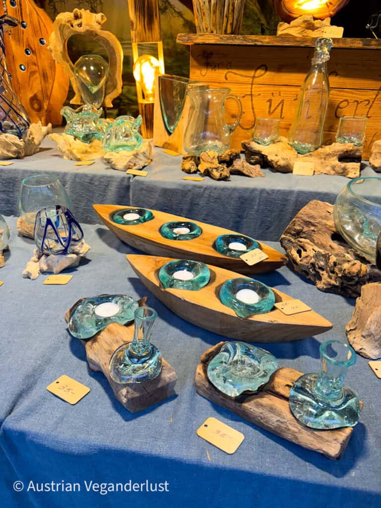 Hand-blown glassware including tealight holders, pitchers, and decorative vases creatively set on wooden stands and blue fabric at a rustic Christmas market stall.