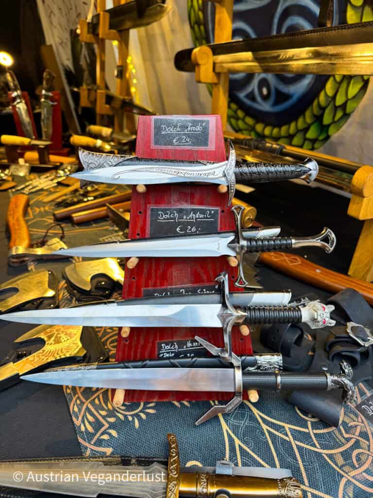 A close-up of ornate replica daggers and short swords, including labels like “Dolch Frodo” and “Dolch Anduril,” displayed vertically at a medieval-themed market stand.