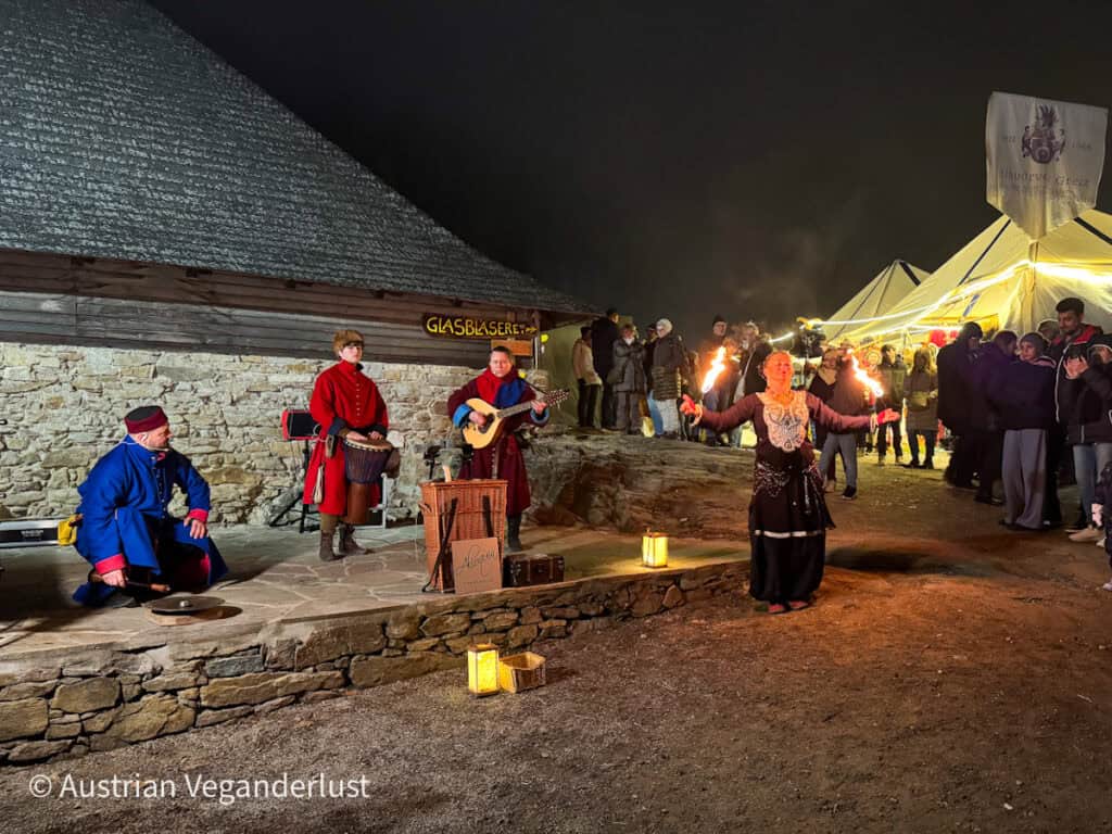 A fire performer entertains a bundled-up crowd at night, backed by medieval-style musicians and illuminated tents at the Aggstein Christmas market.