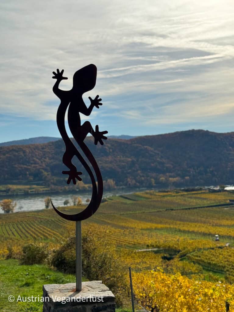 A silhouette sculpture of a gecko mounted on a stone pedestal, set against a backdrop of vineyard-covered hills and the Danube River under a soft, cloud-streaked sky.