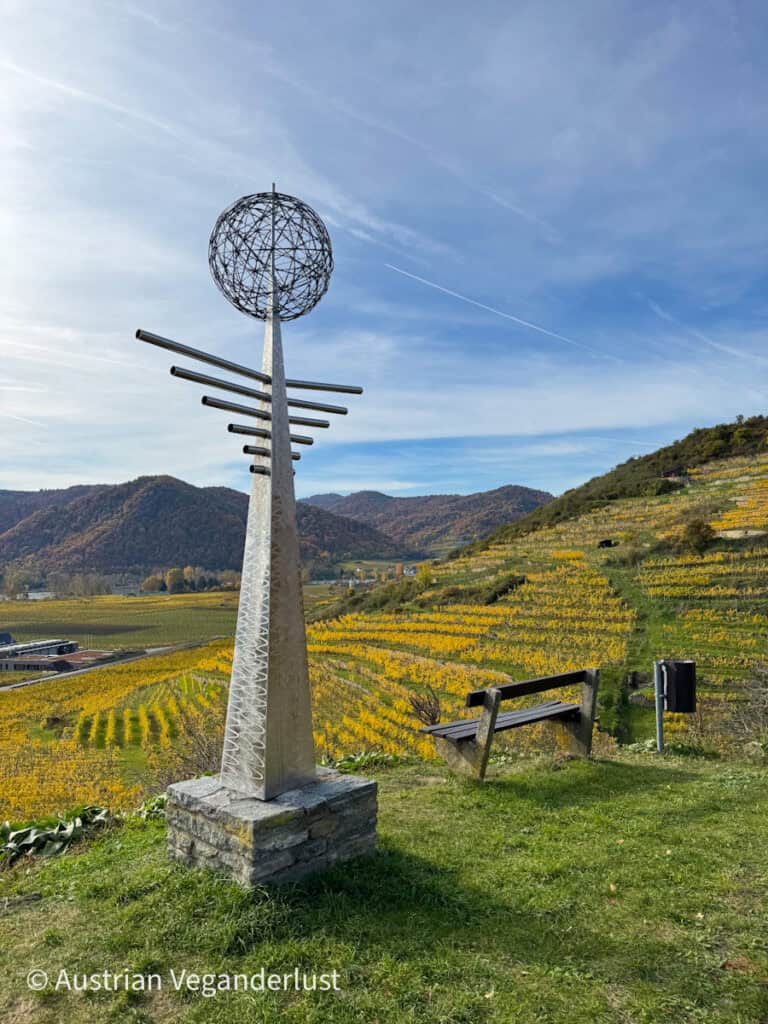 A tall metal sculpture with a spherical wire globe on top and radiating horizontal rods, standing beside a bench overlooking terraced vineyards in the Wachau Valley.