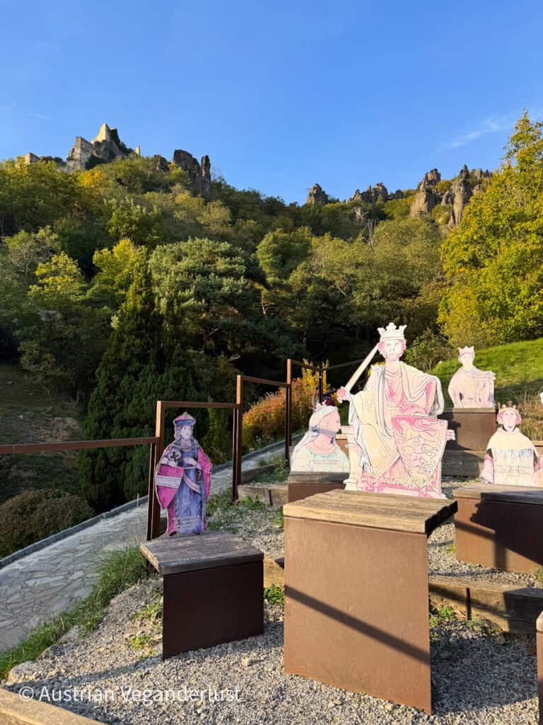 Cutout illustrations of historical figures arranged on wooden platforms, set against a lush green hillside with the ruins of a castle visible at the top under a clear blue sky.
