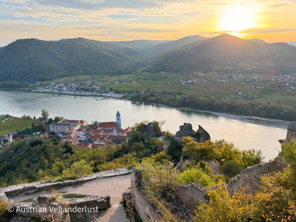 A wide view over the Danube River and the town of Dürnstein at golden hour, seen from the ruins of a hillside castle with layered mountains and vineyards in the distance.