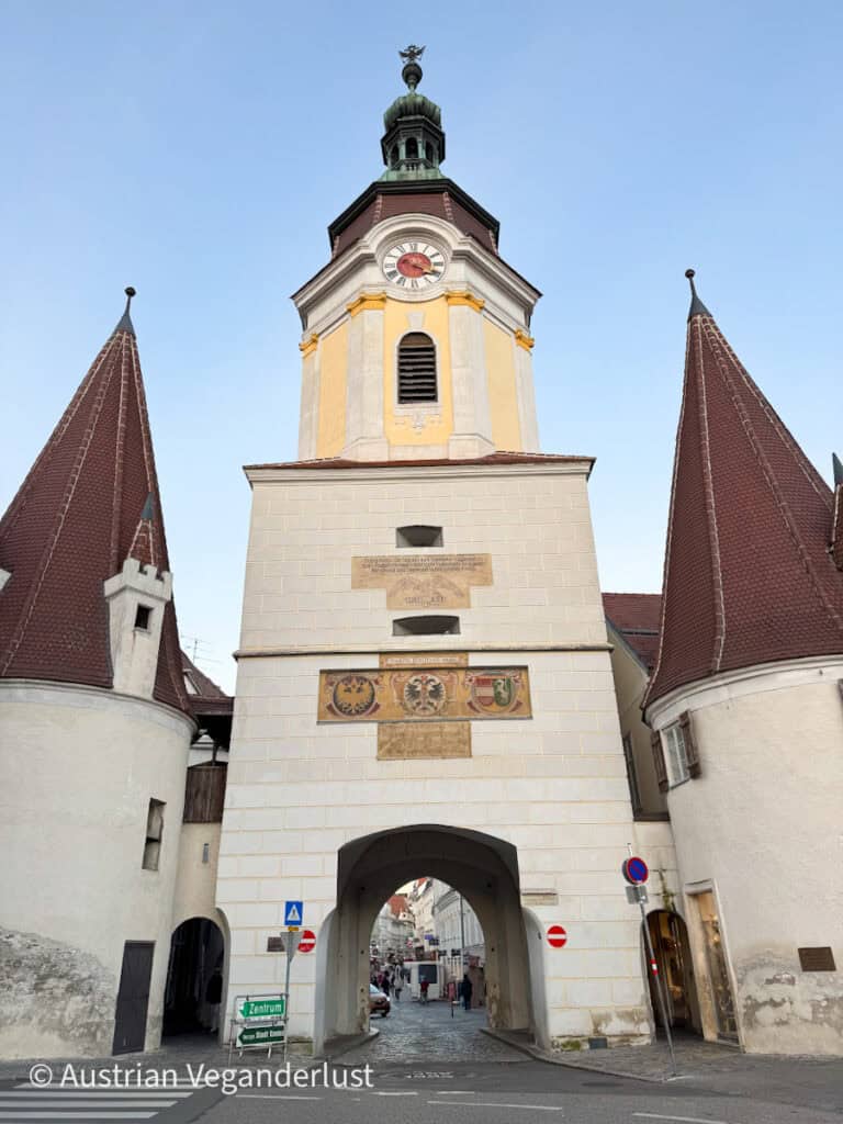 Tall white gatehouse with a clock tower and arched passage leading into the old town, flanked by two conical turrets and decorated with coats of arms and yellow accents.