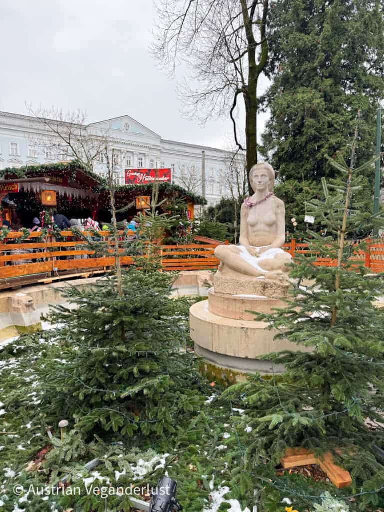 Snow-dusted Christmas trees and a stone statue of a seated woman decorate a quiet garden corner near a cozy Linz Christmas market mulled wine hut.
