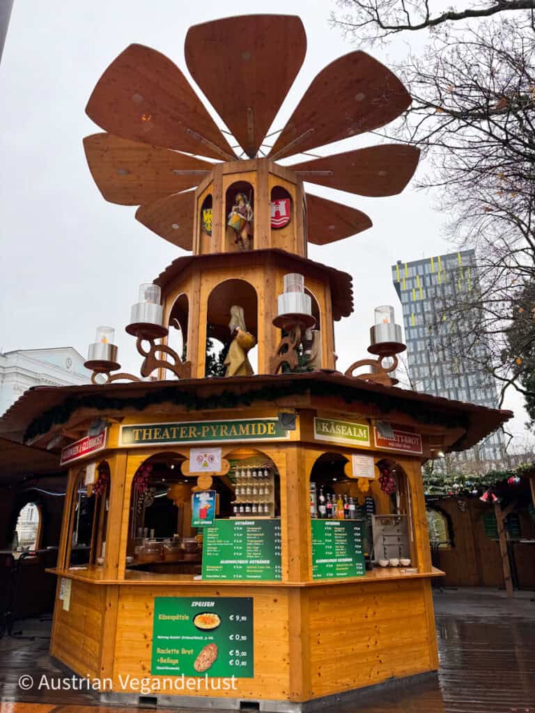 Charming wooden Christmas pyramid-style food stall labeled “Theater-Pyramide” at Linz Christmas markets, serving raclette and Käsespätzle under rotating wooden blades.