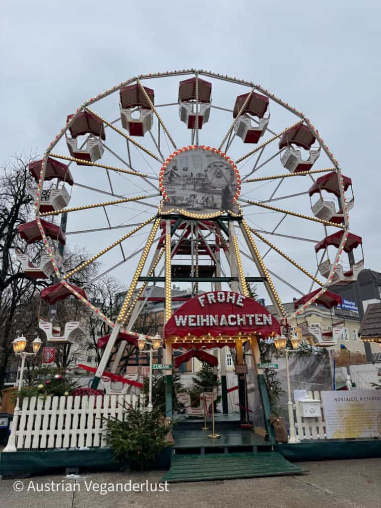 Nostalgic red-and-white Ferris wheel lit with holiday lights and a “Frohe Weihnachten” sign at its entrance, standing tall at the Linz Christmas markets.
