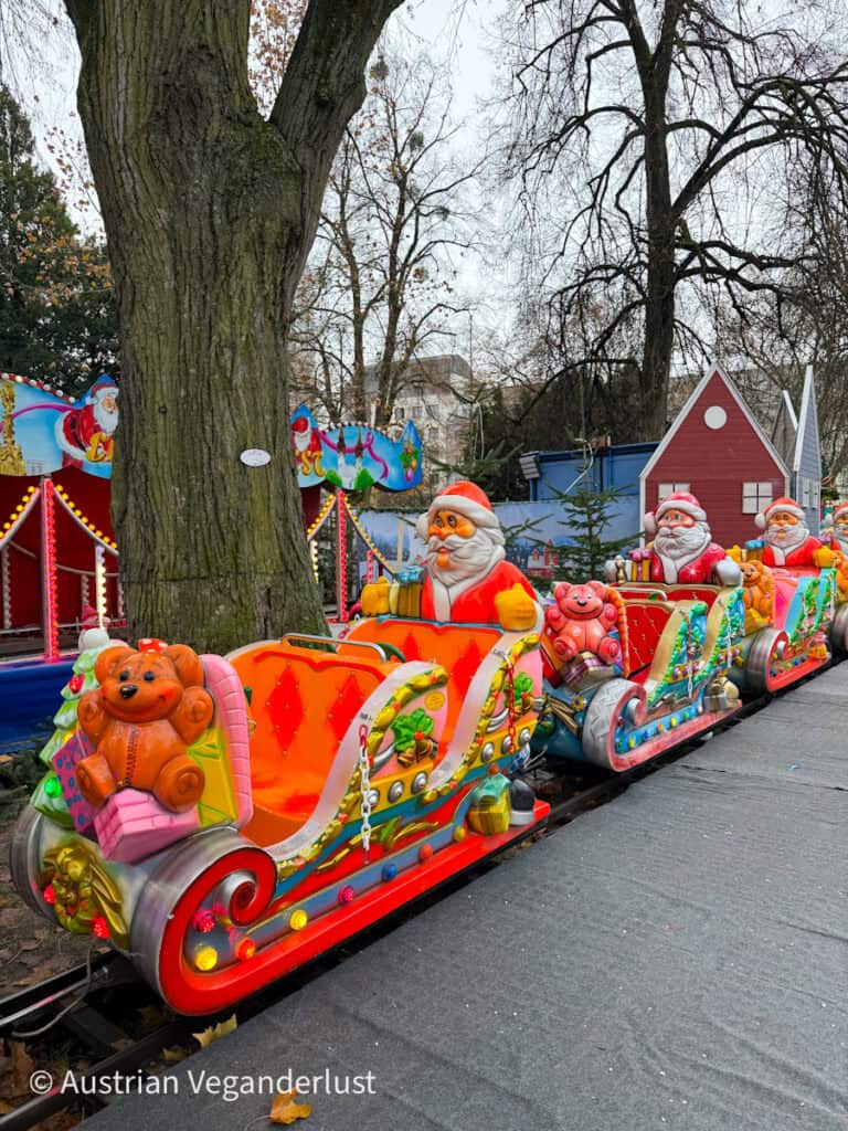 Brightly colored children’s ride at Linz Christmas markets featuring Santa Claus sleigh cars and cheerful teddy bears on tracks winding around large trees.