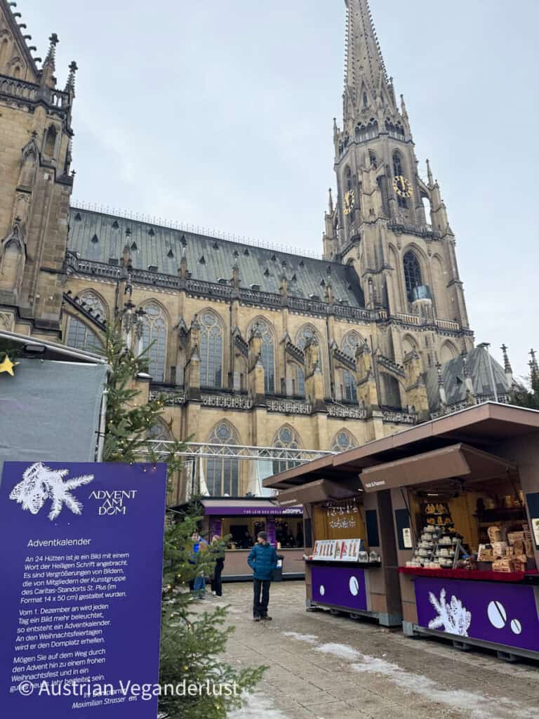 Stalls of the Linz Christmas market set up in front of the towering Gothic New Cathedral, with a large Advent calendar sign and purple-themed booths selling wooden crafts.