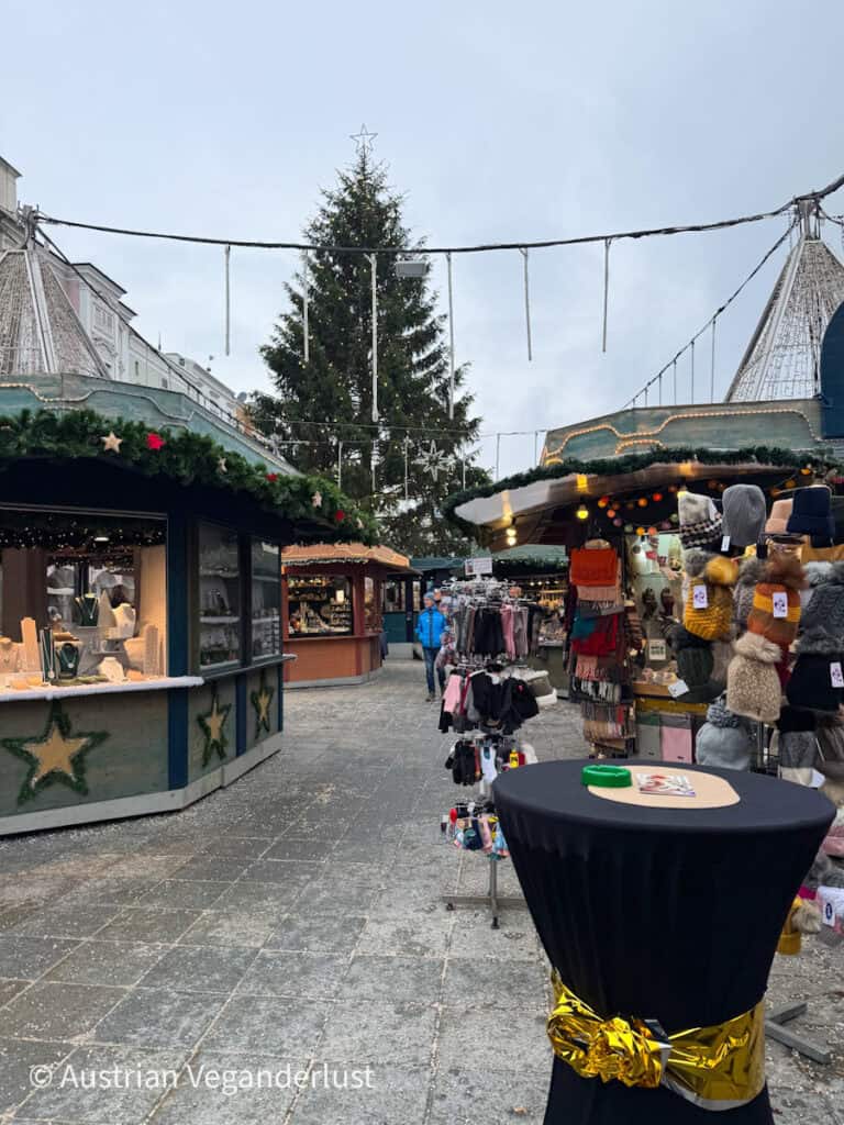 View down a walkway of Linz Christmas markets lined with stalls selling hats, scarves, jewelry, and gifts under festive lights and a towering Christmas tree in the center.