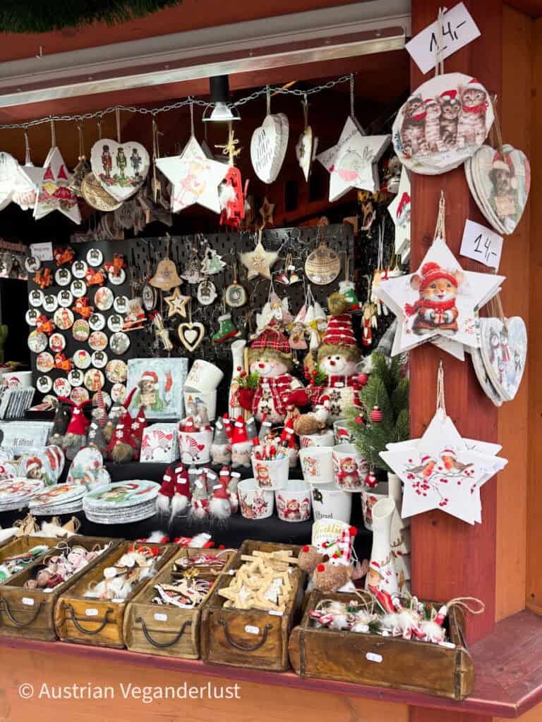 Christmas market stall in Linz brimming with festive decor including wooden stars, Santa mugs, plush elves, and painted ornaments. Prices are displayed in Euros, and shelves are packed with handcrafted holiday gifts.