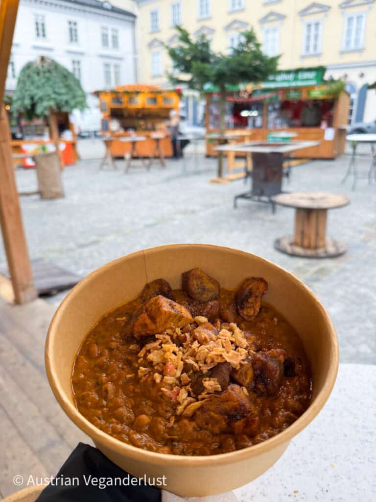 A hearty bowl of plant-based stew topped with crispy onions and roasted plantains, held in front of an outdoor seating area at Linz Christmas markets.