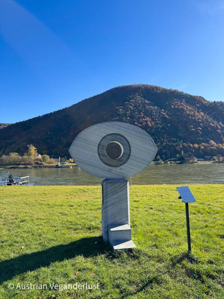 Abstract stone sculpture with a circular eye-like design standing on a grassy riverbank with a forested hill and blue sky in the background.