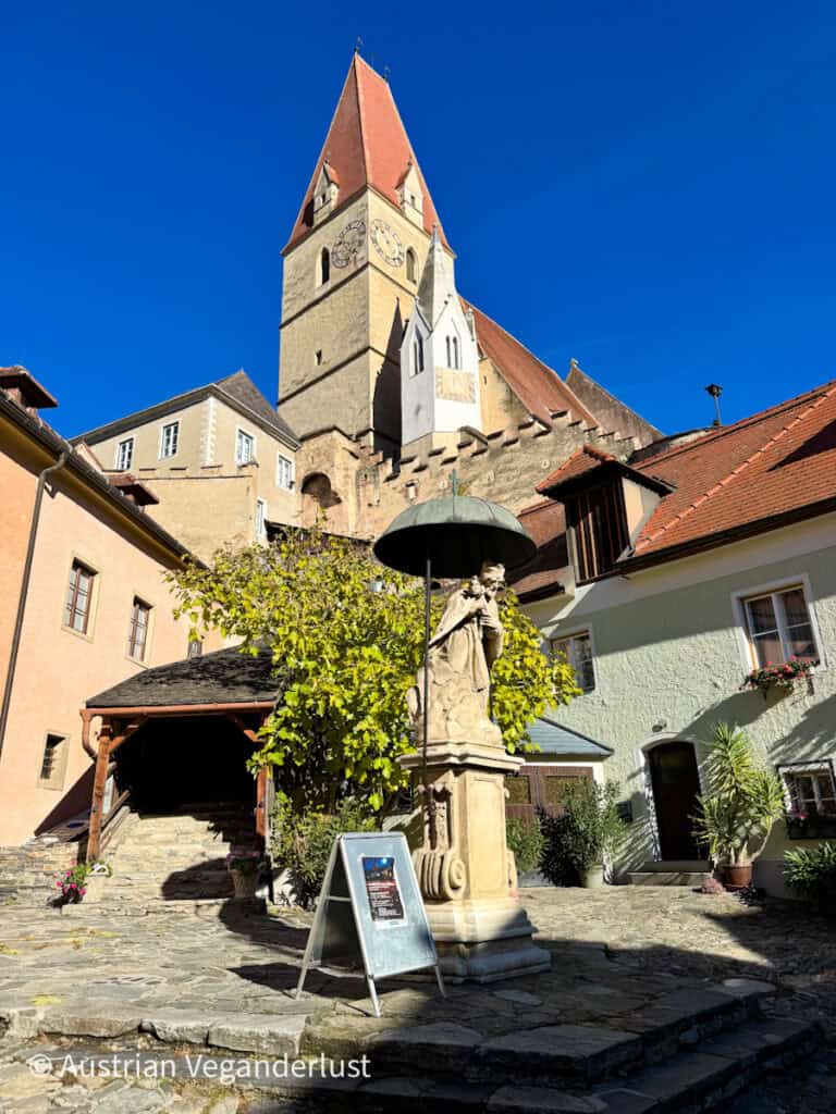Small stone courtyard with a statue under a metal umbrella, informational signboard, and the tall clock tower of a medieval church rising behind surrounding buildings.