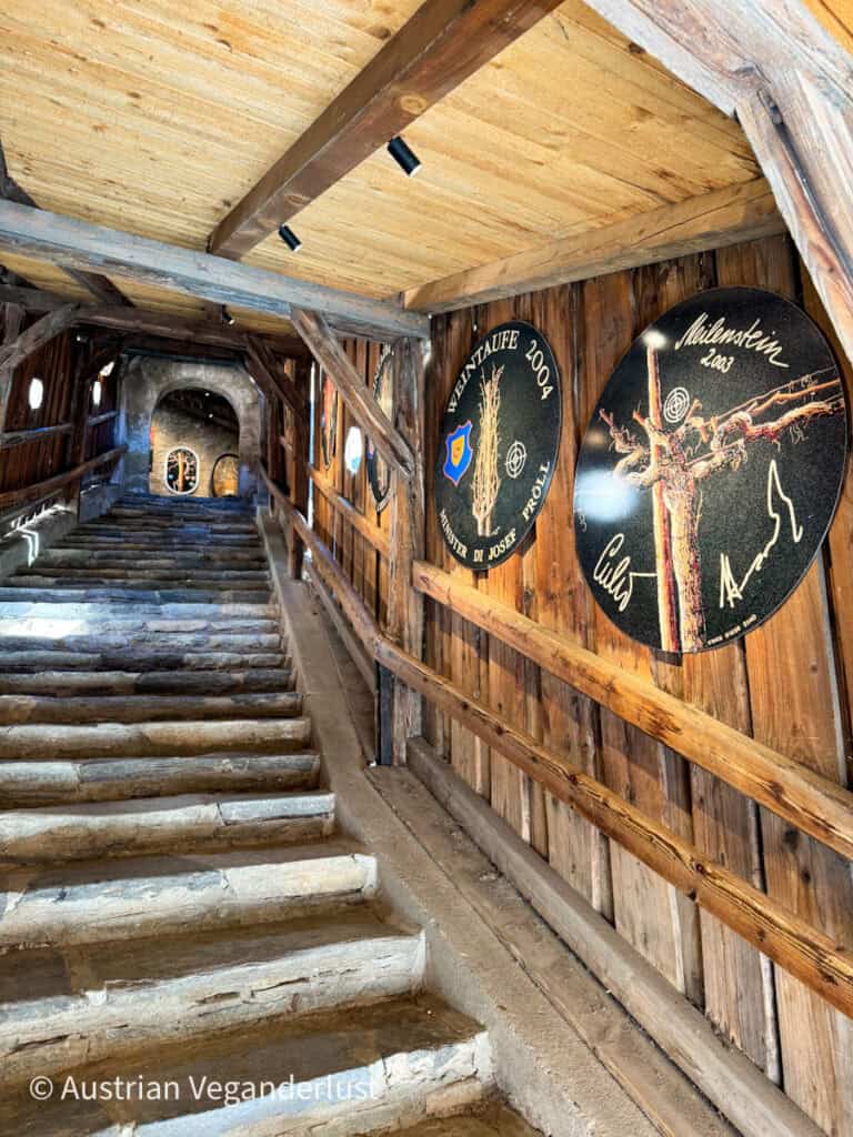 Covered stone staircase inside a wooden passageway, decorated with circular plaques commemorating wine events, leading to a stone archway at the top.