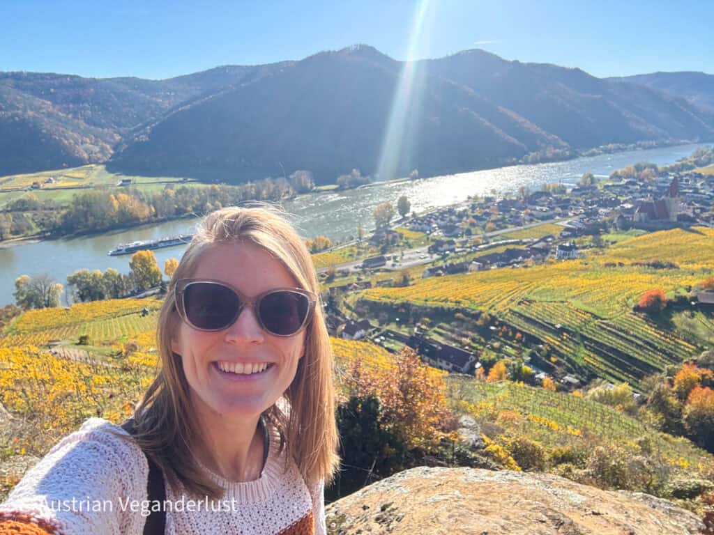 Tina wearing sunglasses and a knitted sweater smiling at the camera with the Wachau Valley in the background. The view overlooks the Danube River, terraced vineyards in golden autumn hues, and a village with a red-roofed church nestled between forested hills.