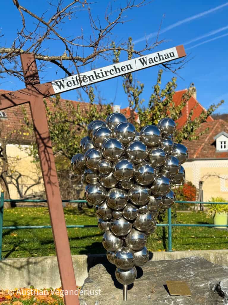 Shiny metal sculpture shaped like a bunch of grapes, placed beneath a sign reading "Weißenkirchen / Wachau" with traditional Austrian houses in the background.