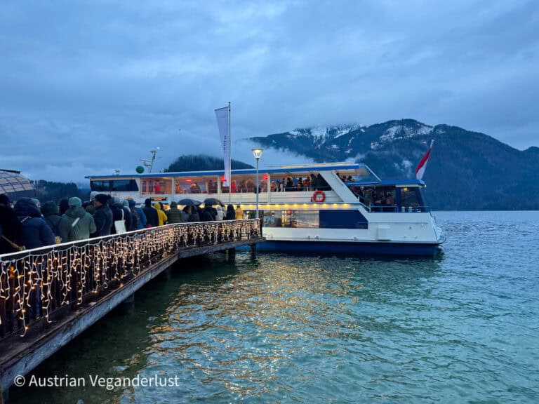 Visitors in warm coats line up on a dock decorated with fairy lights to board a festive boat on Lake Wolfgangsee, with snowy mountain peaks in the background at dusk.