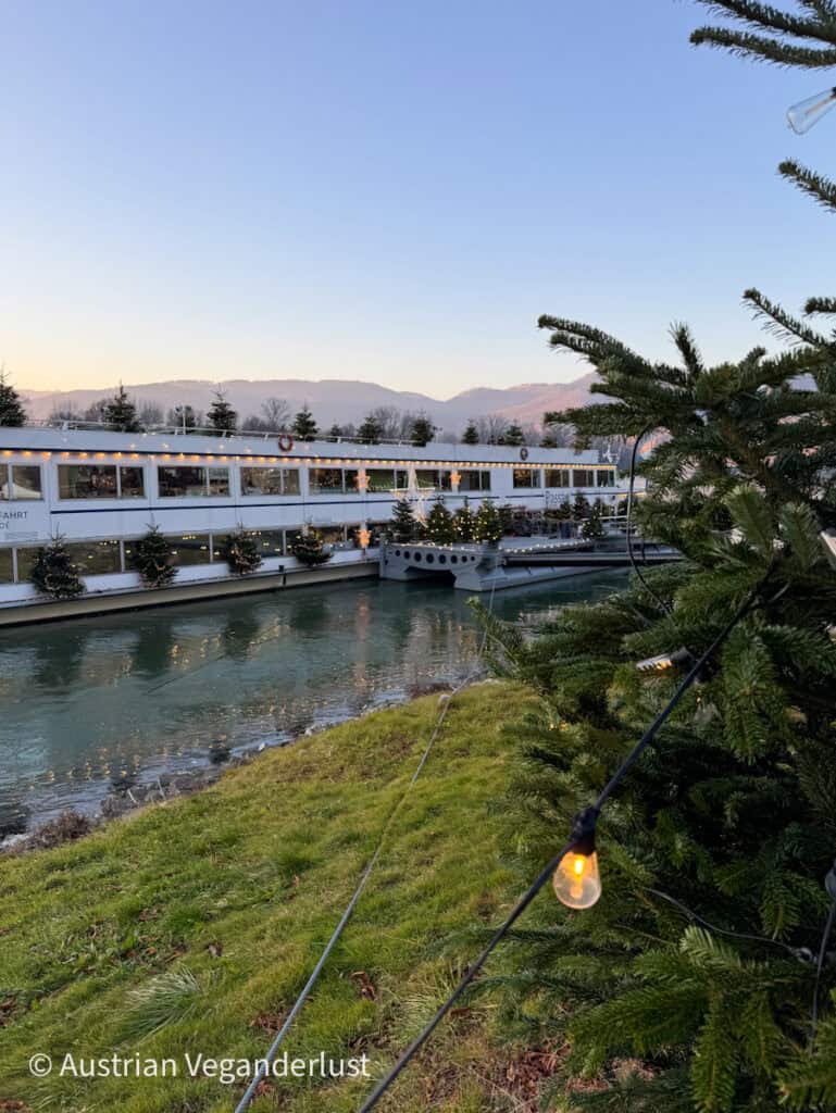 White river cruise ship decorated with Christmas trees and lights, docked on a calm river with mountains in the background at sunset, part of the Dürnstein Christmas markets experience.