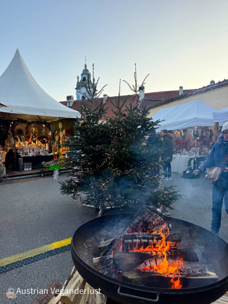 Cozy outdoor scene at the Dürnstein Christmas markets with a fire pit burning logs, surrounded by lit Christmas trees and wooden stalls selling crafts, under a twilight sky with church tower visible.