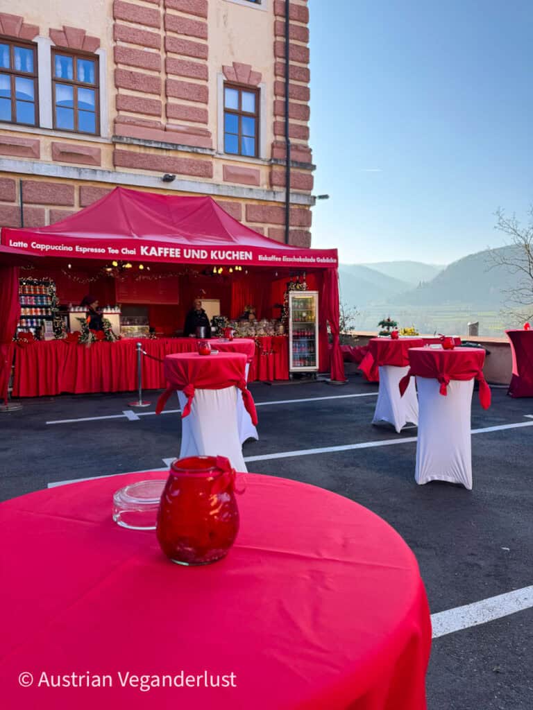 Bright red stall labeled “Kaffee und Kuchen” offering drinks and pastries at the Dürnstein Christmas markets, with tall red-covered tables set against scenic mountain views in the Wachau Valley.
