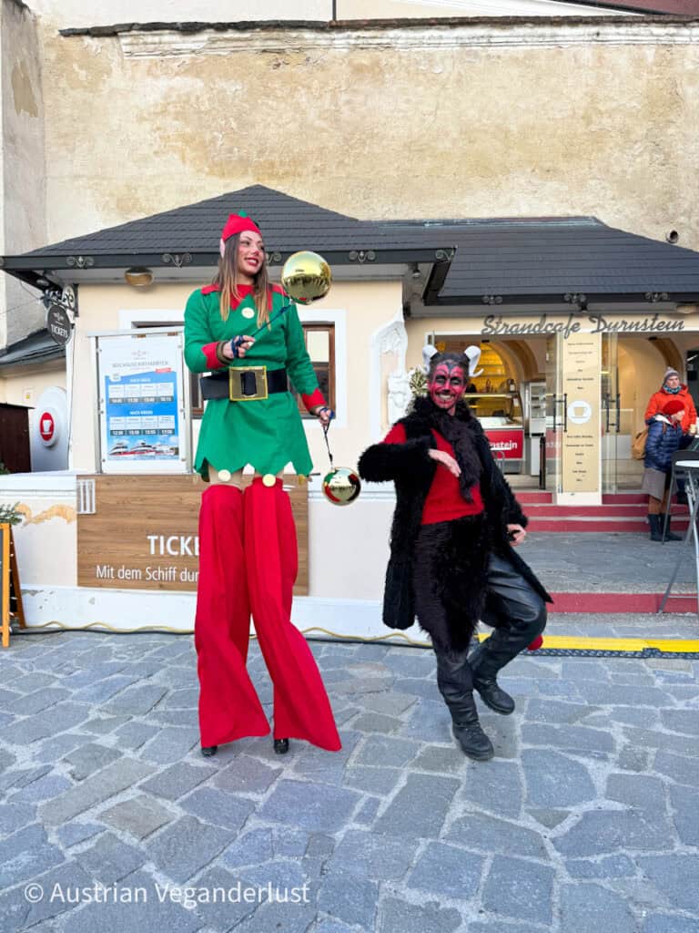 Street performers at the Dürnstein Christmas markets, featuring a smiling woman on stilts dressed as a festive elf and another in a red and black devil costume, entertaining visitors outside the ticket booth.