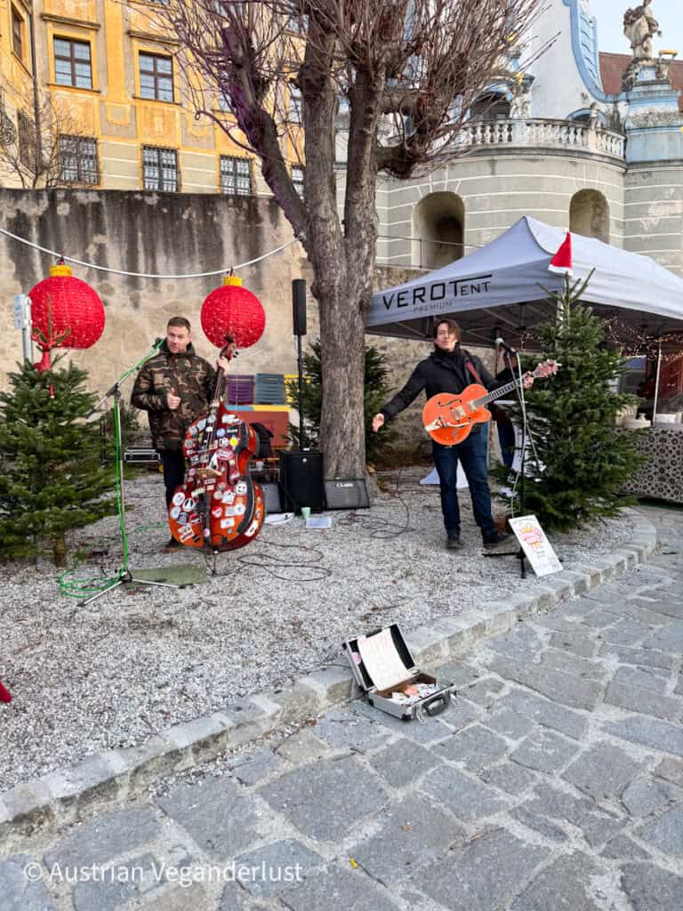 Two musicians performing live next to small Christmas trees under red lanterns in a courtyard near the abbey, creating a lively festive mood at the Dürnstein Christmas markets.