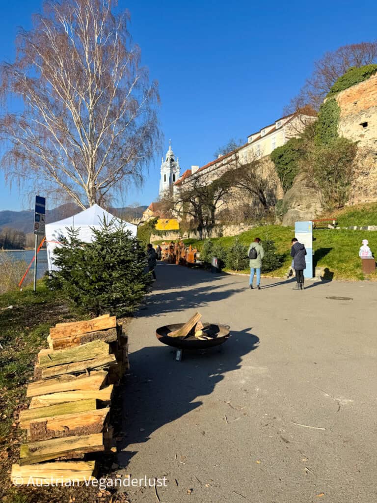 A sunny riverside path near the Dürnstein Christmas markets with a stack of firewood and an unlit fire bowl, framed by the abbey tower and stone walls of the Wachau Valley.