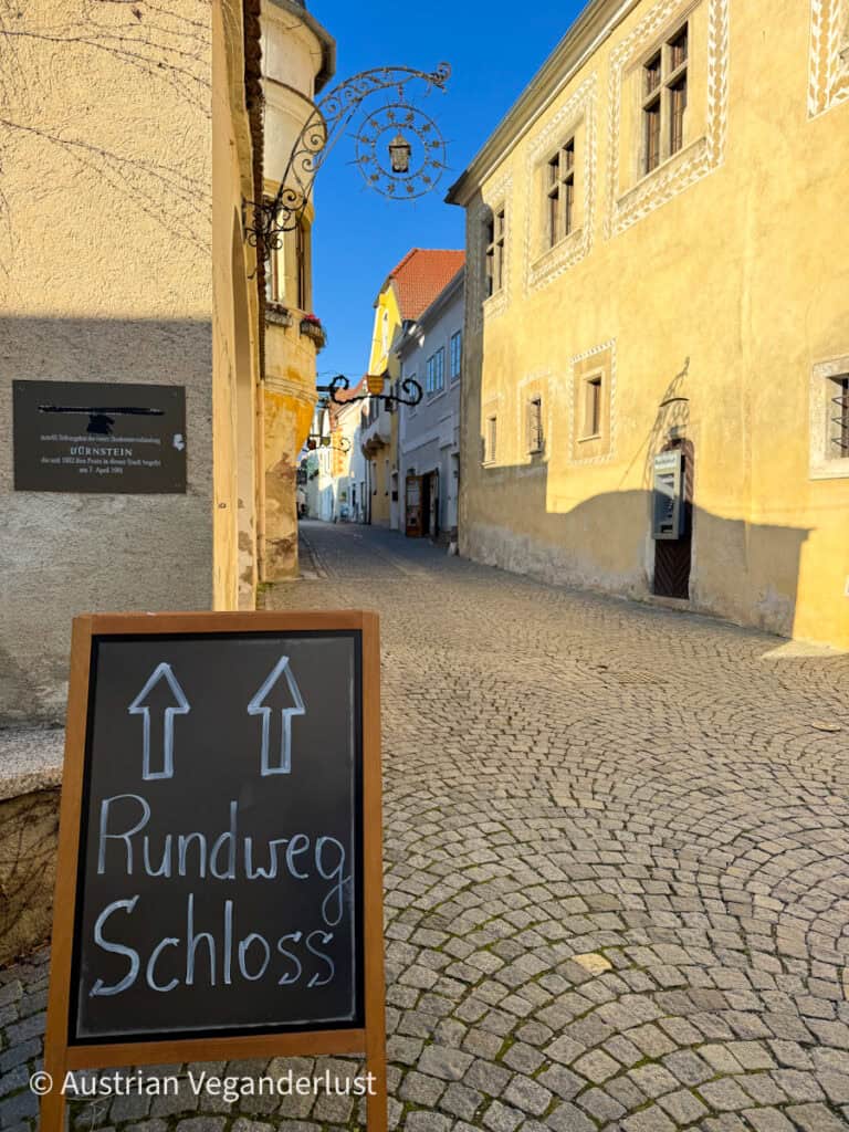 A chalkboard sign reading “Rundweg Schloss” points visitors through a charming cobblestone alleyway in Dürnstein’s old town, guiding them toward the castle path during the festive Christmas season.
