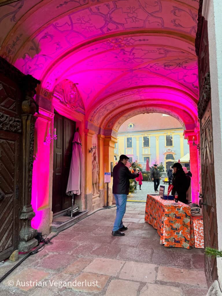 Arched entryway with ornate pink lighting leads into a courtyard at the Dürnstein Christmas markets, where a vendor booth with a bright orange tablecloth welcomes arriving guests.
