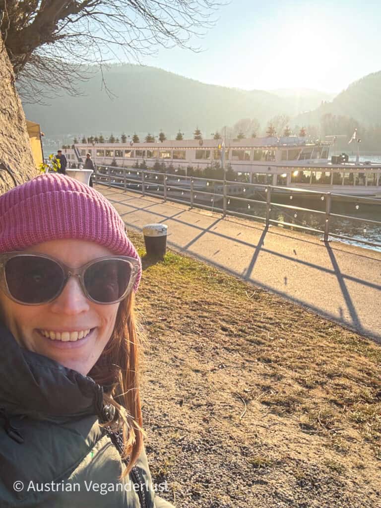 A smiling visitor wearing sunglasses and a pink beanie takes a selfie near the decorated river cruise ship docked for the Dürnstein Christmas markets, with a glowing sun setting behind misty hills.