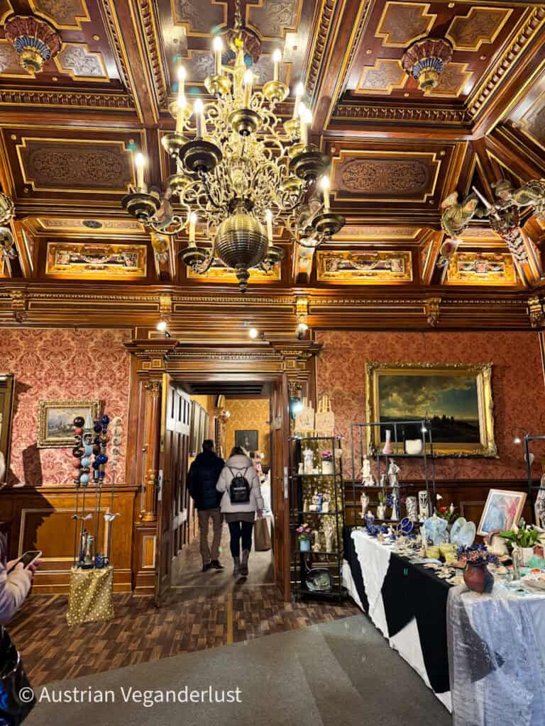 Elegant chandelier and wood-paneled ceiling above Christmas craft stalls inside Grafenegg Castle, showcasing local handmade ceramics and ornaments.