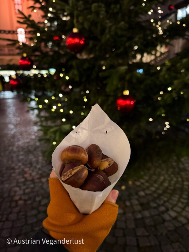 Paper cone filled with roasted chestnuts held in front of a decorated Christmas tree with red baubles at night inside the market courtyard.