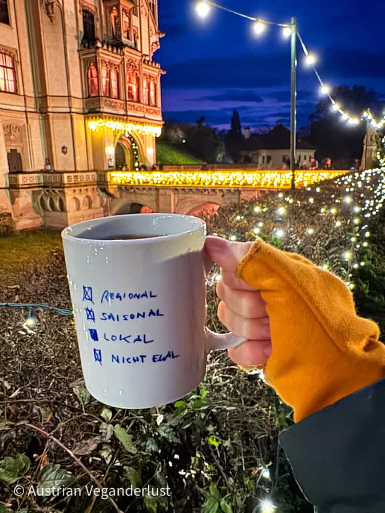 White mug with a handwritten checklist reading “regional, seasonal, local” held in front of the illuminated castle bridge and twinkling lights at the Grafenegg Castle Christmas Market.