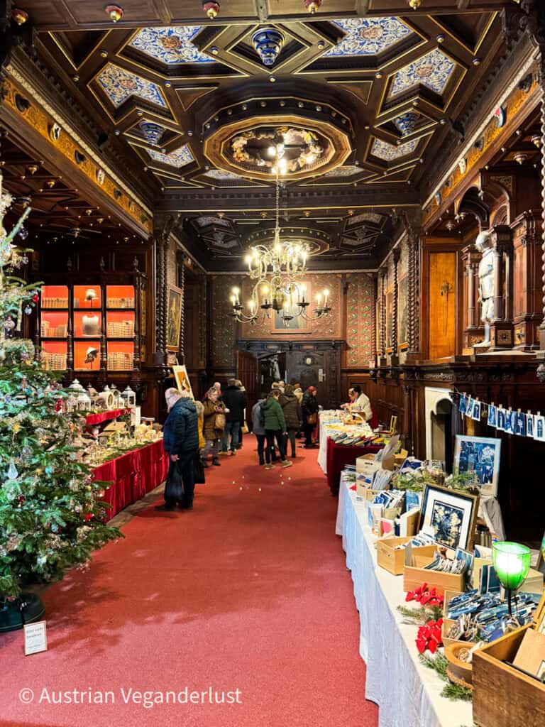 Handmade crafts and postcards for sale inside a richly decorated hall at Grafenegg Castle during the Christmas market, under an ornate ceiling and chandeliers.