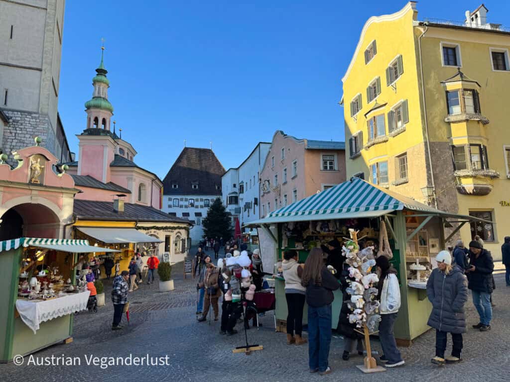 Colorful Christmas market scene in a quaint European town square, with pastel buildings and a green-striped stall selling handmade plush animals. Shoppers browse under a clear blue sky beside a pink baroque-style church and a distant decorated Christmas tree.