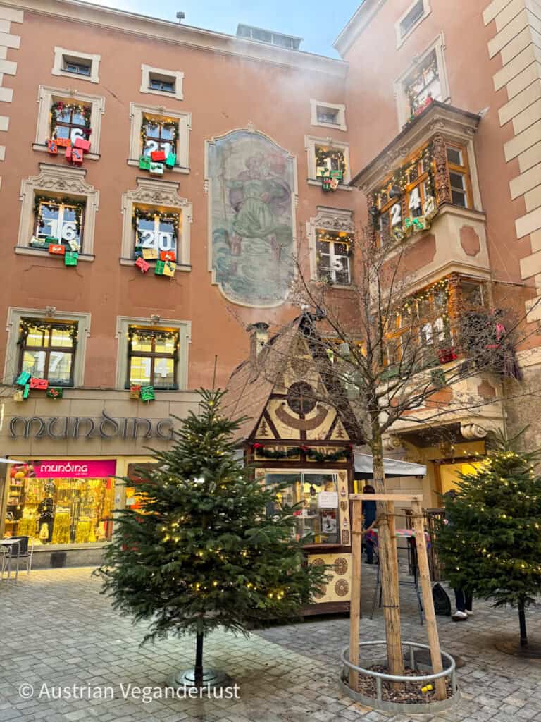 A festive building facade turned into a giant advent calendar with numbered windows and gift decorations, surrounded by small Christmas trees and a fairy-tale themed market stall.