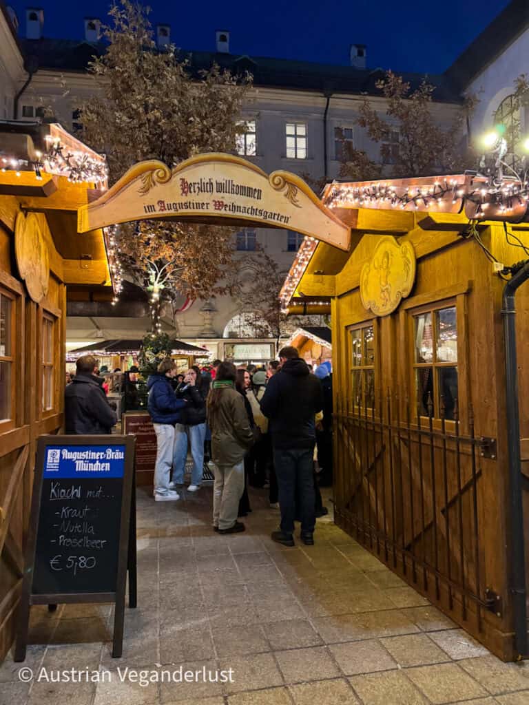 Entrance to the Augustiner Weihnachtsgarten Christmas market in the evening, with a warmly lit wooden arch reading "Herzlich willkommen im Augustiner Weihnachtsgarten" and a crowd gathered under string lights. A chalkboard menu offers Kiachl with kraut, Nutella, or Preiselbeer for €5.80.