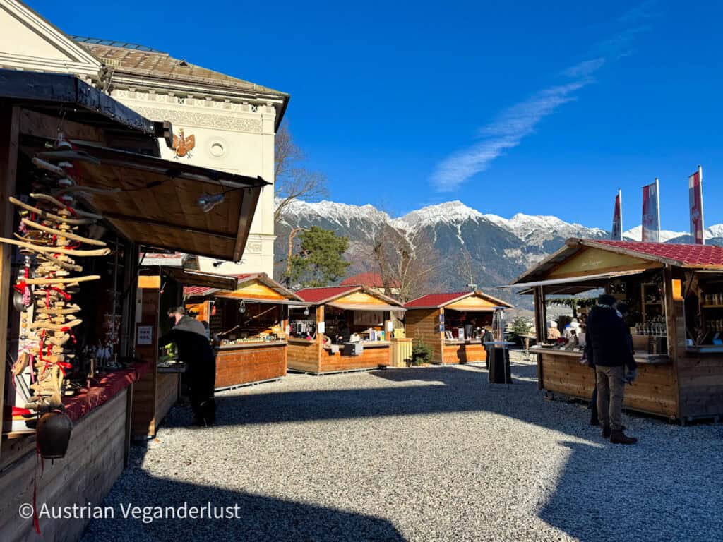 A quiet daytime Christmas market with wooden huts selling goods, set against a bright blue sky and snow-capped mountain range. Few visitors browse the stalls in the open gravel courtyard.