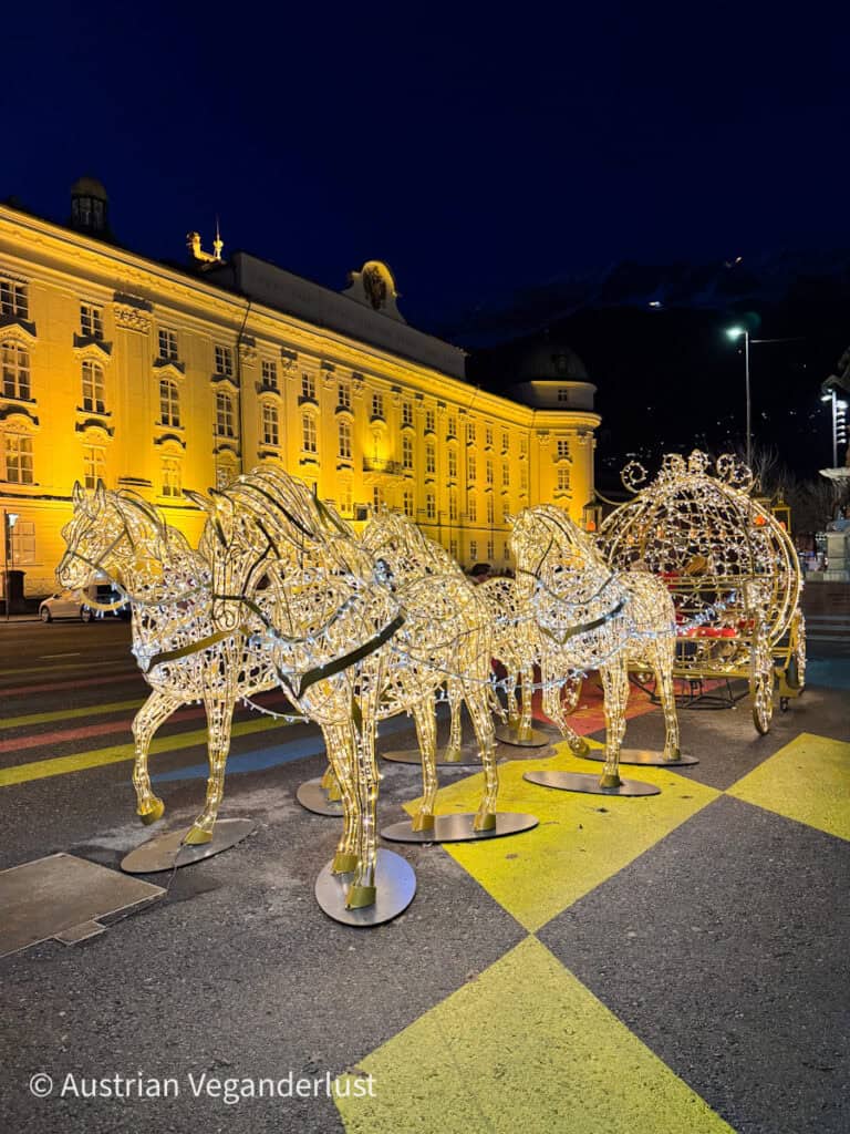 Illuminated horse-drawn carriage Christmas display glowing in front of the Hofburg Imperial Palace in Innsbruck at night. The golden lights highlight the ornate baroque architecture while snow-covered mountains fade into the background.