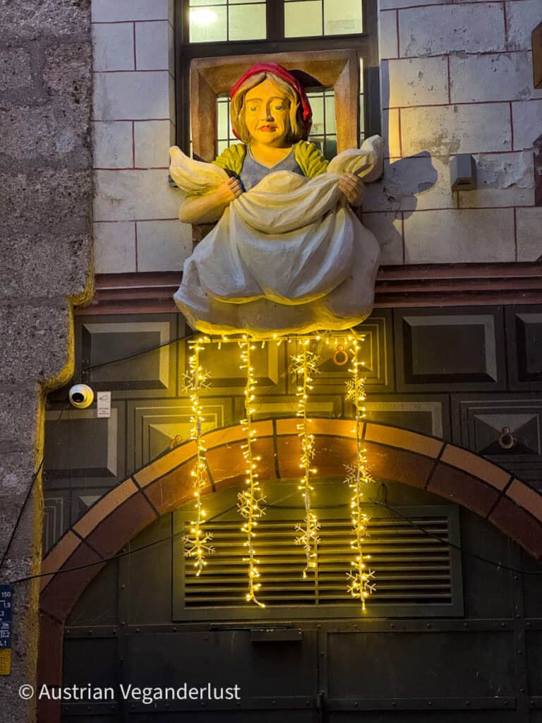 A sculpted figure of Frau Holle leans out of a window shaking a sheet, symbolizing falling snow, with glowing snowflake string lights hanging below. The display is set on a traditional Innsbruck building during the Christmas season.