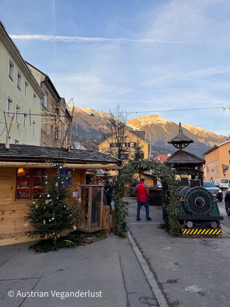 A small-town Christmas market with a decorated archway and mini train engine in front of snow-capped mountains. Wooden stalls and festive trees line the quiet street.
