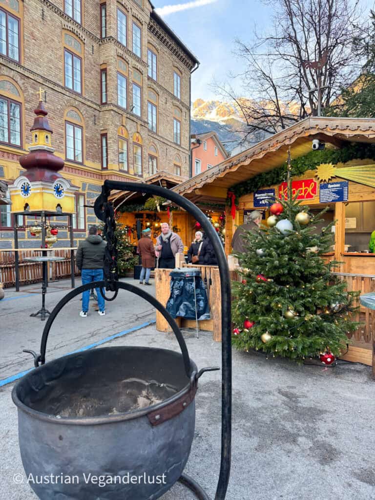 Close-up of a rustic iron cauldron with ash inside, set in front of a festive Christmas stall and decorated tree. A historic building and mountains glow in the late afternoon light.
