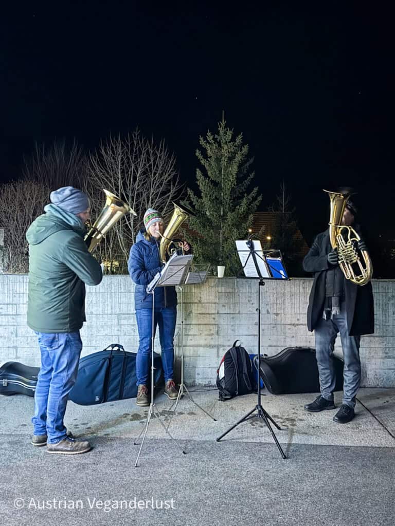 Three musicians play brass instruments outdoors at night in winter attire, illuminated against a concrete wall with bare trees and a pine in the background.