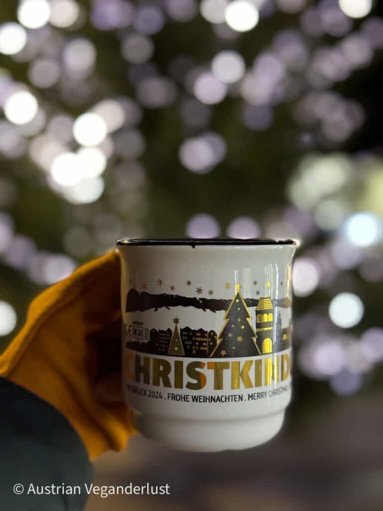 A hand in a yellow glove holds a souvenir mug with golden illustrations and the word “Christkind” in bold letters. The blurred lights of a Christmas tree glow softly in the background.