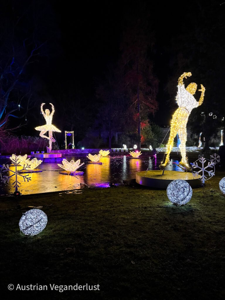 Light sculptures of a figure skater and ballerina pose beside a pond with glowing lotus flowers and oversized snowflakes in vibrant winter colors.