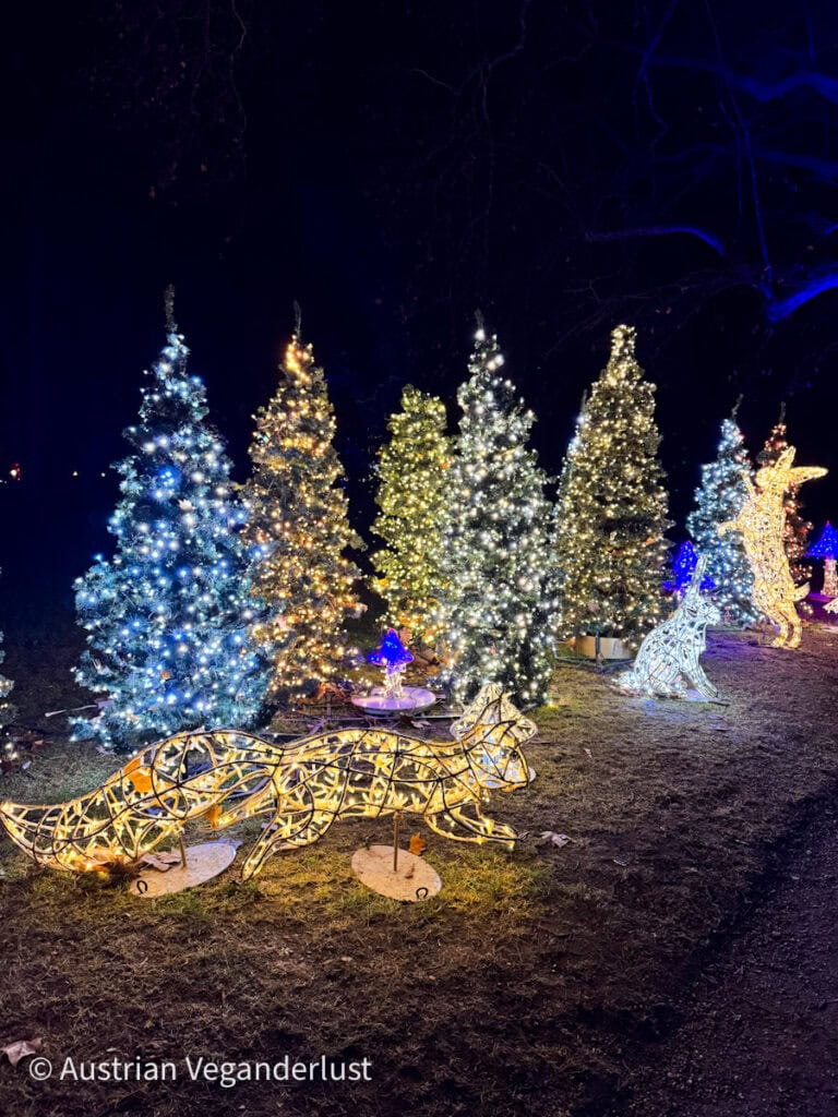 A line of brightly lit Christmas trees in various colors with glowing animal sculptures, including a fox and deer, creating a magical woodland scene.