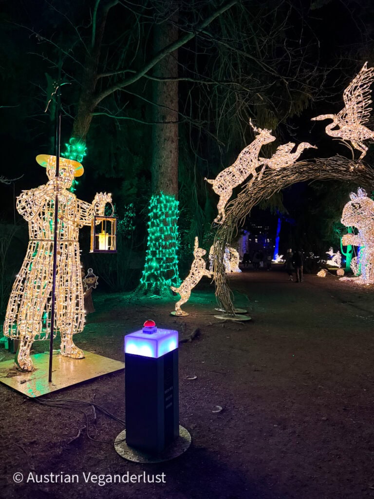 Archway of glowing animal lights featuring deer, rabbits, and a heron, with a lantern-holding figure nearby and a button pedestal at the front of the enchanted Lumagica trail.