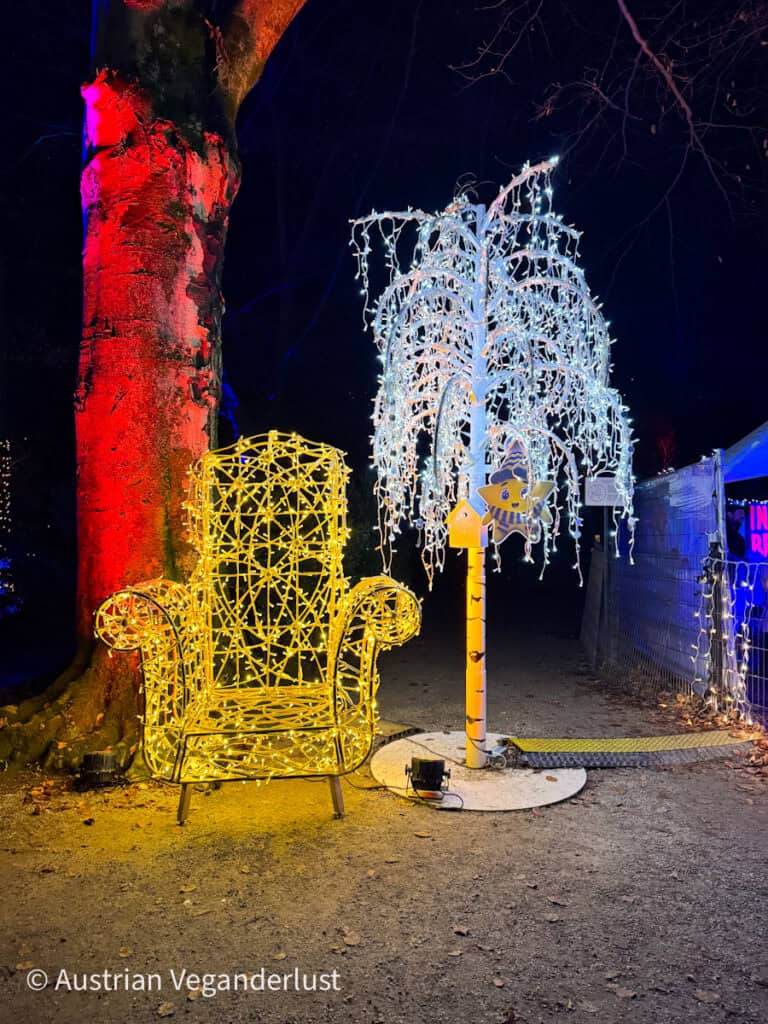 A glowing yellow light sculpture of an oversized armchair sits beside a white weeping tree light display, marking a whimsical rest area along the Lumagica trail.